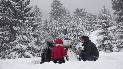 Family building a snowman in park, lovely child fixing buttons, snowed trees, beautiful winter family portrait, real teamwork