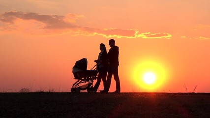Fototapeta premium Mother and father silhouette walking together with baby pram, close colored sky, sunset sun shinning, conceptual