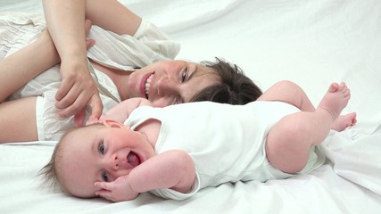 Obraz na płótnie Canvas Portrait of attractive young mother and lovely baby with blue eyes, smiling face, blue, laying down on bed and holding hands, pure love