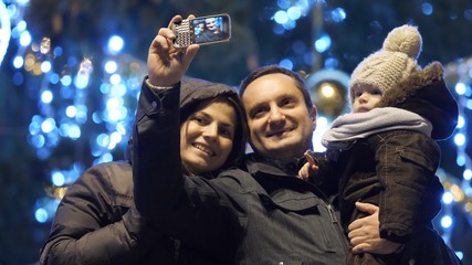 Young parents and baby taking selfie pictures in front of Christmas tree outdoor
