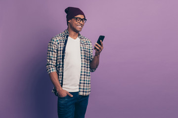 Fototapeta na wymiar Portrait of cheerful positive millennial with beaming toothy smile using holding modern gadget device in hands putting palm in denim trousers isolated violet background
