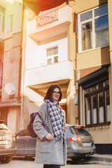Attractive positive young girl wearing glasses in a coat on the background of buildings on cars