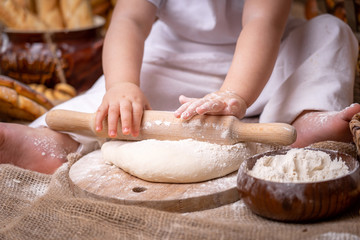 Obraz na płótnie Canvas a small child sits on a wooden table and rolls out the dough with a rolling pin,flour is scattered around and bread lies. the child develops fine motoriku gets skills