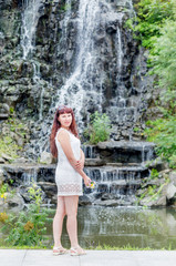 Portrait of cute young caucasian woman in nice white dress in the summer park. Beautiful waterfall on background.