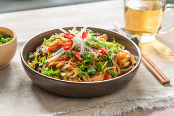 Asian vegan lunch - spicy chow mein noodles with carrots, pepper and spinach with sliced radish, spring onions and chili peppers on a rustic table