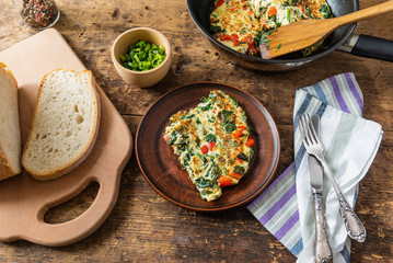 A piece of frittata with spinach and red pepper on a plate and a cutting board with bread, rustic style, top view