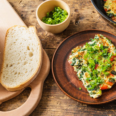 Homemade frittata with spinach and red pepper, sprinkled with spring onions on a plate and cutting board with bread, rustic style - Italian healthy breakfast
