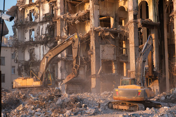 Fototapeta premium Building demolition with hydraulic excavator. Dismantle of destructed house ruins at bright sunsest