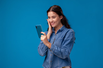 Young brunette woman is posing with a smartphone standing on blue background.