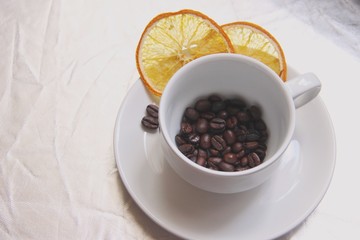 One White Cup and saucer filled with roasted coffee beans, with two slices of dried orange and a pair of coffee beans on the silver background. Pair Cup and saucer, coffee beans, slices of dried