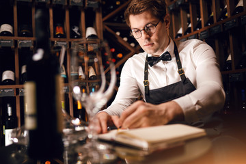 Thoughtful male sommelier looking at a very wide range of wines from the main regions of the world placed of wine shelves, writing in note pad some things working at wine restaurant.