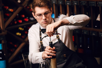 Elegant young sommelier with bow tie uncorking bottle of wine in wine boutique. Wine tasting social...