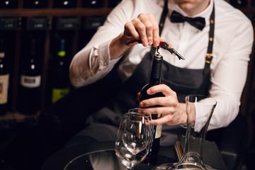 Elegant young sommelier with bow tie uncorking bottle of wine in wine boutique. Wine tasting social...
