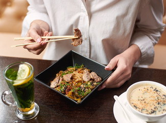 Woman ordering oriental style meal in restaurant. Client enjoying lunch including soup with seeds, warm salad with meat and lemon mint tea. Close up hands with manicure holding food sticks.