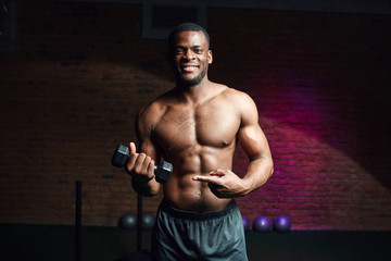 Fototapeta na wymiar Fitness portrait of dark-skinned shirtless male bodybuilder with perfect six pack pointing at dumbbell in his hand. prompting to sport and training.