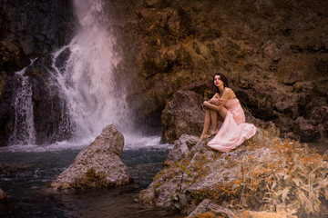 Attractive girl posing on the rock among in fairy fast forest in autumn and beside beautiful waterfall with blue water. Fairy tale.