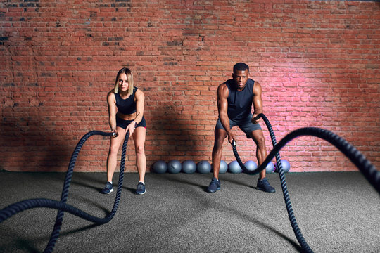 Multiethnic fitness couple training cardio workout together, exercising with battle ropes at indoor gym.