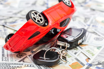  Overturned red car toy with car key on background of dollar bills. Car accident.