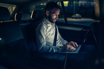 Businessman in car working on laptop in back seat.