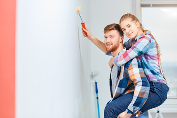 Cheerful bearded man giving piggyback ride to his wife as she paints wall with a brush, while renovating the apartment