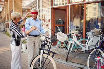 elderly woman covering eyes of senior man for surprise with gift bicycle.