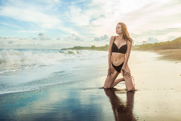 Slim sexy brunette girl in a bathing suit kneeling on the beach.