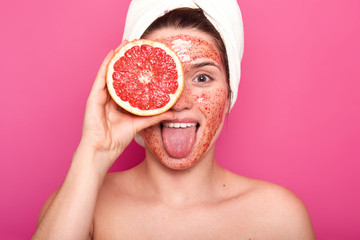 Beautiful young woman with bright scrub on her face holds half of rape grapefruit in one hand, sticks out her tongue, having white towel on her head, looks fresh and delighted. Skin care concept.