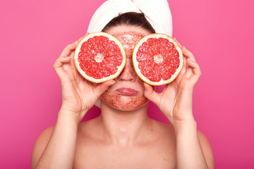 Dissatisfied naked woman frowns her face holding parts of rape grapefruit in both hands, having colourful face scrub and white towel. Unsmiling model poses over bright pink background in studio.