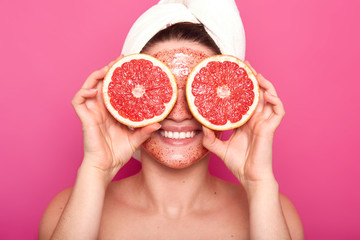 Close up portrait of positive attractive girl smiling sincerely, holding two parts of grapefruit, covering her eyes with fruite, having satisfied facial expression, being in good mood, face scrub.