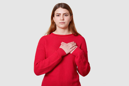 Calm dark haired female with serious facial expression, keeps both palms on heart, being touched by something, dressed in casual red sweater, isolated over white background. Body language concept