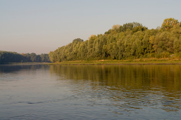 Wide river flowing across green forest. Fall. Reflections of sun in the water