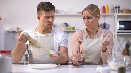 Clumsy man stretching dough, looking at wife, couple has difficulty making cake