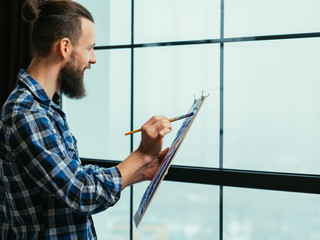 Fine art. Creative process. Handsome bearded guy inspired by view outside the window, enjoying painting.
