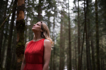 Model wearing an elegant red dress walking into a dark and gloomy forest while looking up at the sky