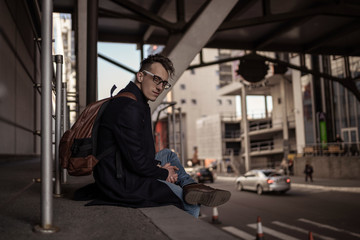 Young man in black coat and glasses with backpack sitting on city street near shopping center. Street style photo shoot.