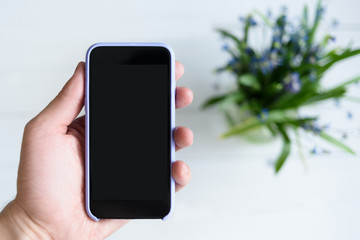 Male hand with a smartphone. Black blank screen. Table with flowers on background