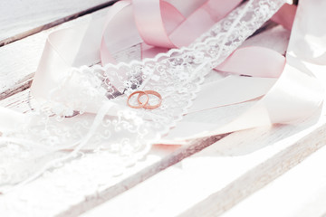 Gold wedding rings with pink lace and ribbons lie on a white wooden surface.