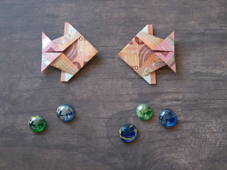 Two origami fish made of ten euro notes facing each other on dark wooden table area
