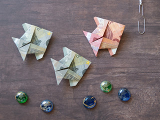 Three origami fish made of ten and five euro notes swimming towards fishing-rod on dark wooden...