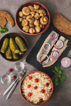 Traditional Russian appetizer and vodka, sauerkraut with cranberries, herring, pickled cucumbers, pickled mushrooms and rye bread on a brown background. Top view, copy space.