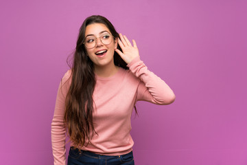 Teenager girl over purple wall listening to something by putting hand on the ear