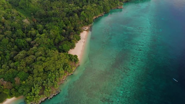 Aerial: flying over tropical Hatta island white sand beach Banda Islands Maluku Indonesia lush green forest turquoise water coral reef sunset sky