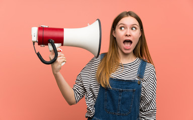Naklejka premium Young woman with overalls over pink wall taking a megaphone that makes a lot of noise