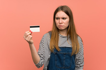 Young woman with overalls over pink wall taking a credit card without money