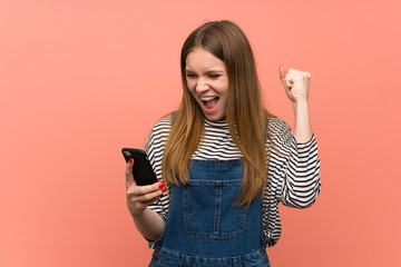 Young woman with overalls over pink wall celebrating a victory with a mobile