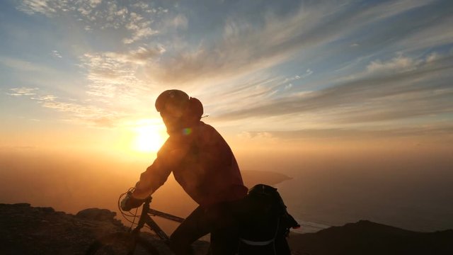 Man rides a bicycle high in mountains near cliff edge above the ocean against beautiful dramatic sunset background.