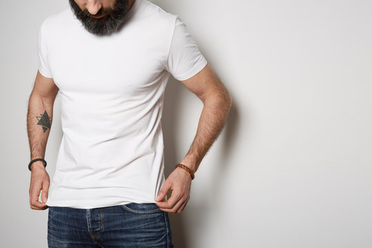Attractive brutal tattooed bearded guy poses in blue jeans and blank gray t-shirt premium summer cotton, on white background. Mockup Copy Paste Advertisement