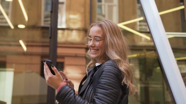 Cheerful caucasian girl in stylish glasses walking on the street in sunny day and texting on the smart phone. Outdoors, urban