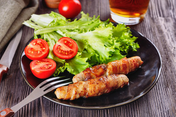 Appetizing sausages wrapped in bacon and grilled next to tomatoes, lettuce and and glass of beer are on a wooden table