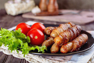 Appetizing sausages wrapped in bacon and grilled next to tomatoes and lettuce are on a wooden table